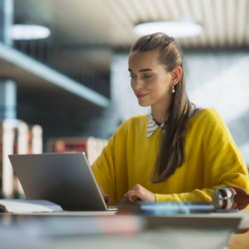 A young professional woman in a bright yellow sweater looking at an open laptop with a confident smile in a modern office or library setting.